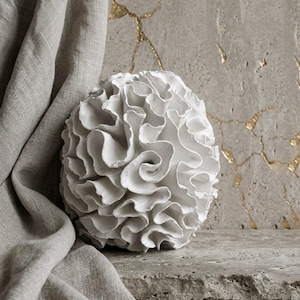 Pode incluir: Um objeto decorativo branco, esférico, com uma superfície texturizada e com babados. O objeto é colocado sobre uma superfície de pedra, com um tecido cinza drapeado no lado esquerdo. O fundo apresenta uma parede texturizada com detalhes dourados.