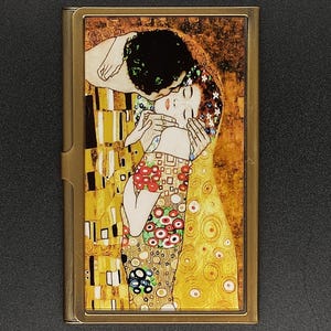Puede incluir: Una imagen enmarcada en latón de una pintura que representa a una pareja abrazándose. La pintura está en un estilo que recuerda a "El Beso" de Gustav Klimt con un fondo dorado y detalles coloridos.