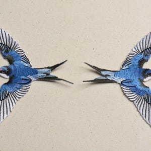 Op de afbeelding: Twee gestikte zwaluw-patches in blauw en wit. De vogels kijken in tegengestelde richtingen met hun vleugels uitgespreid.