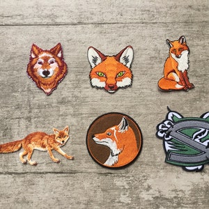Puede incluir: Un conjunto de seis parches bordados con diseños de animales, incluyendo zorros y un lobo. Los parches presentan tonos de naranja, marrón y blanco, con un parche verde y gris con la letra "S". Ideales para ropa o accesorios.