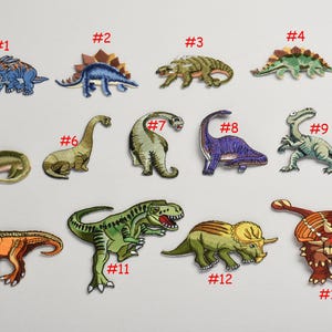 May include: A set of 13 embroidered iron-on patches featuring different dinosaur designs. The patches are colorful and detailed, with a variety of colors including green, blue, red, orange, and purple. The patches are numbered 1 through 13.