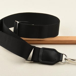 May include: A black adjustable bag strap with silver-tone metal hardware. The strap is made of a durable, woven material and features a leather accent. The strap has a swivel clasp for easy attachment to a bag.