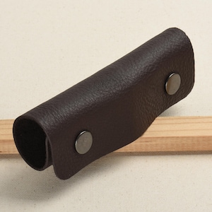 Op de afbeelding: Een bruine leren sleutelhanger met twee metalen drukknopen. De sleutelhanger is opgerold en heeft een gestructureerd oppervlak.