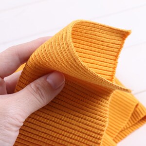 May include: Close-up of a yellow ribbed knit fabric. The fabric is folded in half and held in a hand.