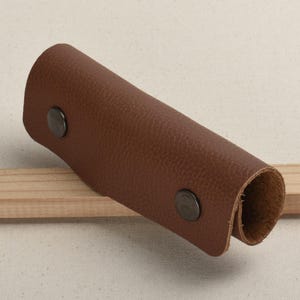 May include: A brown leather handle with two metal rivets. The handle is rolled at the edges and has a textured surface.