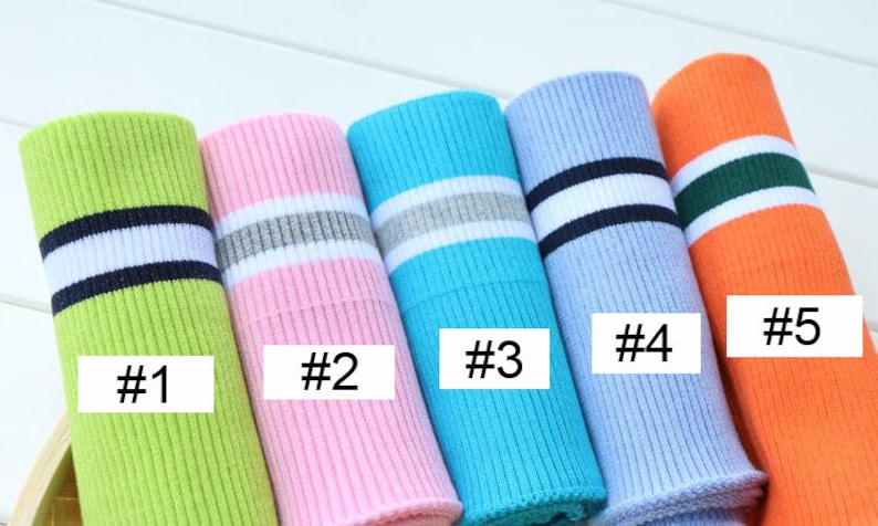 Op de afbeelding: Vijf verschillende soorten geribbelde breisels met contrasterende strepen. De strepen zijn in verschillende kleuren, waaronder wit, zwart, grijs, groen en blauw. De stof is opgerold en gelabeld met de nummers 1 tot en met 5.