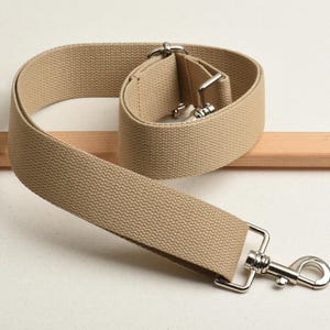 以下が含まれることがあります： 銀色の金属製の留め具が付いたベージュのキャンバスストラップ。ストラップは半分に折り畳まれ、留め具が見えています。