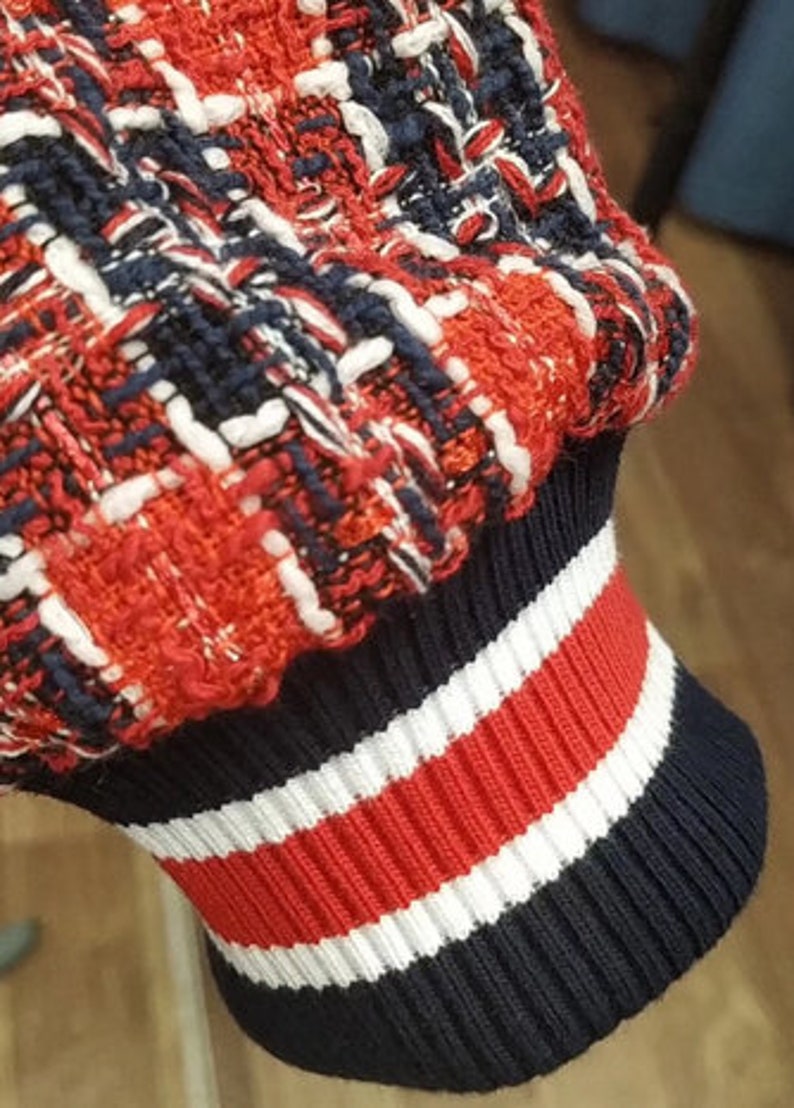 Op de afbeelding: Close-up van een rood, wit en blauwe gebreide manchet met een textuurpatroon. De manchet is gemaakt van een zachte, rekbare stof en heeft een geribbeld ontwerp.