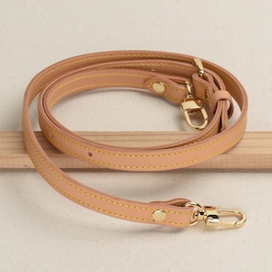 Puede incluir: Una correa de cuero marrón claro con herrajes dorados y costuras. La correa está doblada por la mitad y tiene dos cierres giratorios.