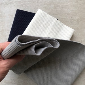 K&ouml;nnte beinhalten: Drei Rollen geripptes elastisches Gewebe in Schwarz, Wei&szlig; und Grau. Das Gewebe ist gefaltet und wird in einer Hand gehalten.