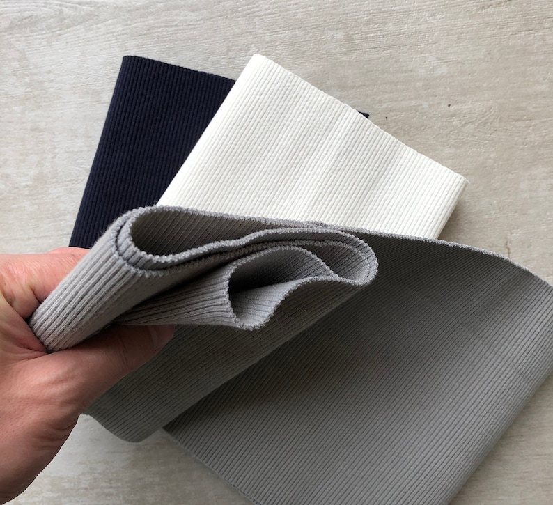 K&ouml;nnte beinhalten: Drei Rollen geripptes elastisches Gewebe in Schwarz, Wei&szlig; und Grau. Das Gewebe ist gefaltet und wird in einer Hand gehalten.