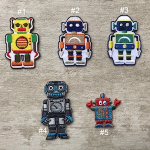 Könnte beinhalten: Fünf bunte Roboter-Aufnäher mit gestickten Details. Die Roboter sind in verschiedenen Farben wie Gelb, Blau, Grün und Rot gehalten, mit schwarzen Umrissen und weißem Hintergrund. Ideal zum Aufnähen auf Kleidung oder Accessoires.
