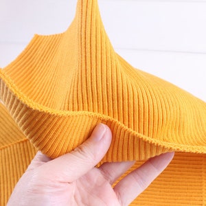 May include: Close-up of a yellow ribbed knit fabric. The fabric is folded in half and held by a hand.