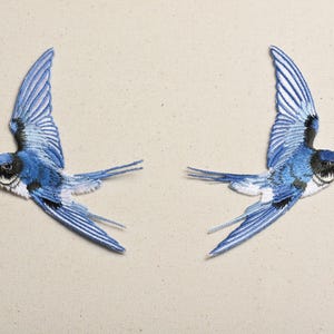 May include: Two embroidered patches of blue swallows in flight. The birds have detailed wings, with shades of blue and white. The swallows have black and white markings on their faces and a touch of pink on their heads. The patches are on a beige background.