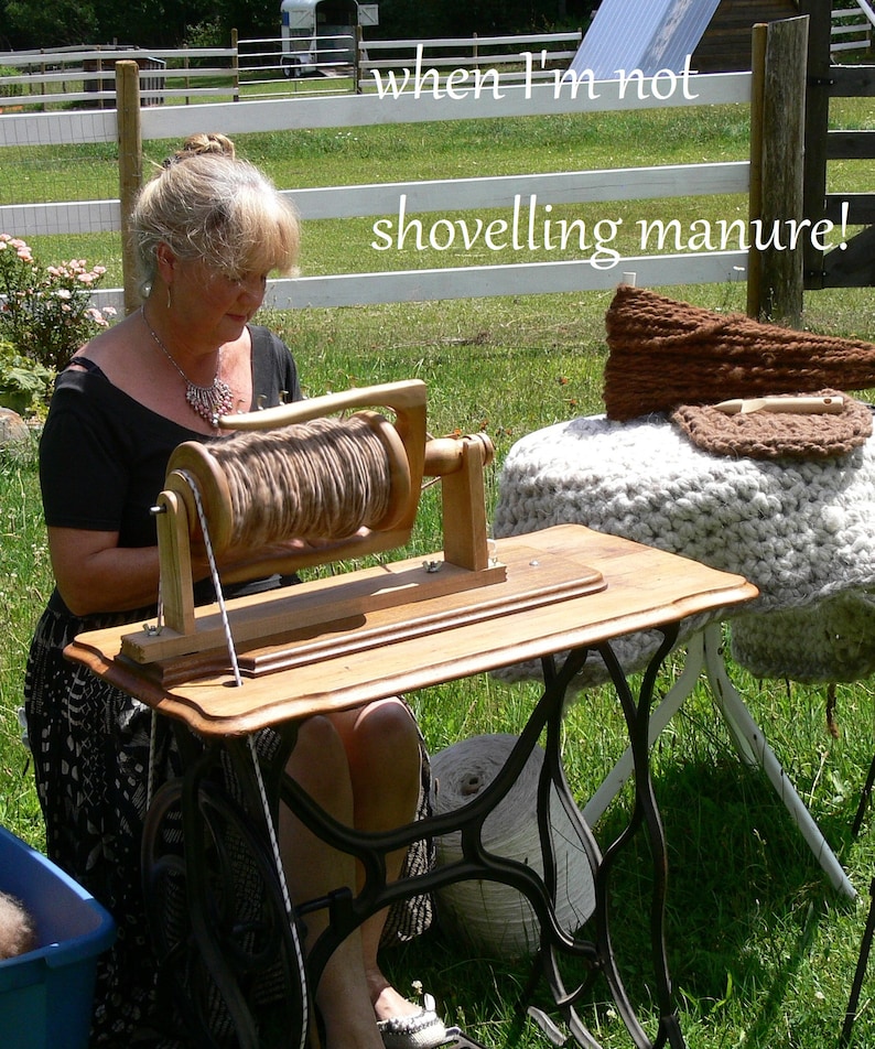 May include: A woman sits at a wooden table with a spinning wheel, winding yarn. The table is a repurposed sewing machine base. The caption reads "when I'm not shovelling manure!"
