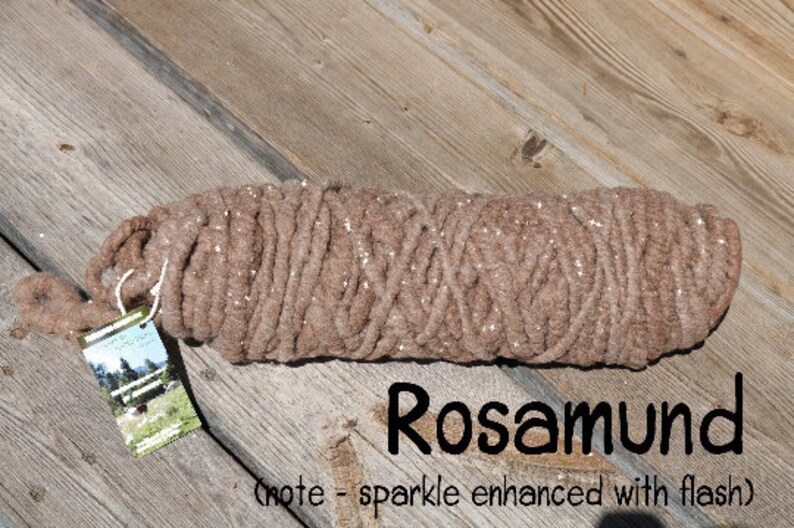May include: A brown yarn with a sparkly finish, rolled up and tied with a white string. The yarn is labeled "Rosamund" and has a note that says "sparkle enhanced with flash".