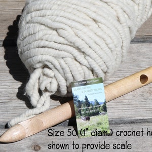 May include: A large ball of white yarn with a 1 inch diameter crochet hook for scale. The hook is made of wood and has a hole in the handle. The yarn is thick and fluffy. The text on the card reads "The Llama Sanctuary, Llama Safari & Guided Tours".