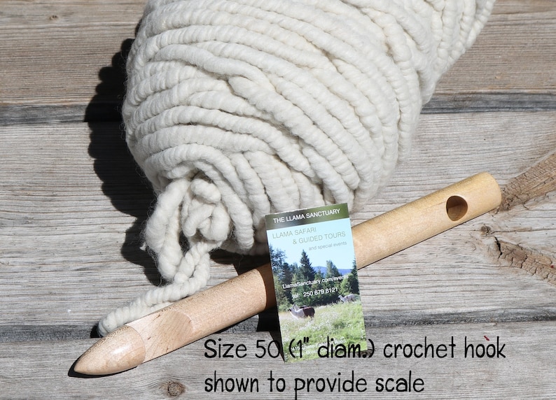 May include: A large ball of white yarn with a 1 inch diameter crochet hook for scale. The hook is made of wood and has a hole in the handle. The yarn is thick and fluffy. The text on the card reads "The Llama Sanctuary, Llama Safari & Guided Tours".