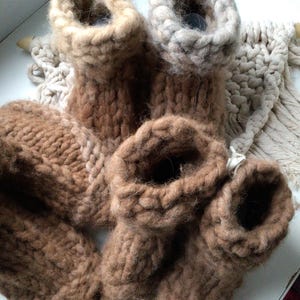May include: A collection of chunky knit booties in various shades of brown and beige. The footwear features a thick, textured yarn construction with a high-top design and a visible ribbed cuff. The items are arranged together in a cluster.