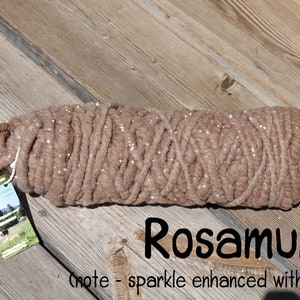 May include: A thick, light brown yarn skein with a subtle sparkle effect, featuring a rectangular product tag with a landscape image and the text Rosamund (note - sparkle enhanced with flash) printed in black on the lower right side.