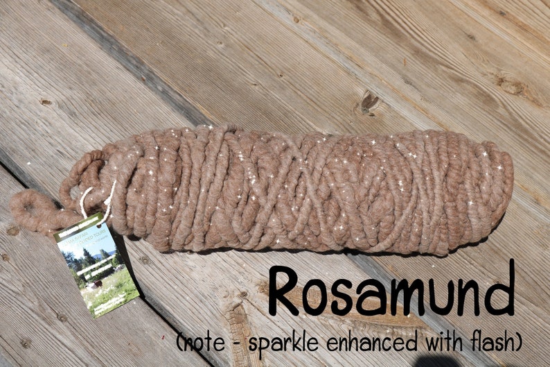May include: A thick, light brown yarn skein with a subtle sparkle effect, featuring a rectangular product tag with a landscape image and the text Rosamund (note - sparkle enhanced with flash) printed in black on the lower right side.