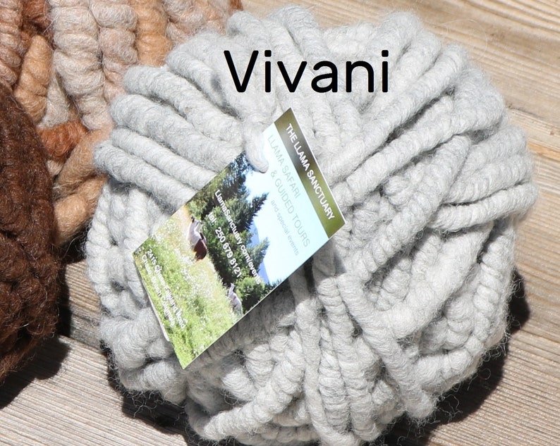 May include: A ball of light gray chunky yarn with a tag labeled Vivani and The Llama Sanctuary Llama Safari & Guided Tours. The yarn is thick and textured, featuring a soft, fibrous appearance. A portion of a brown yarn ball is visible on the left.