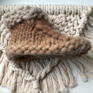 May include: A chunky knit slipper boot featuring a gradient of tan and brown yarn with a thick, textured cuff. The boot is constructed with large, visible stitches and a soft, fuzzy appearance, showcasing a high-top silhouette with a rounded toe.