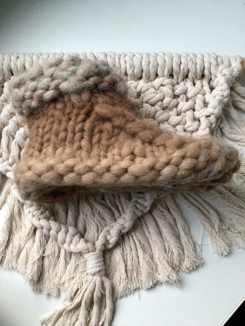May include: A chunky knit slipper boot featuring a gradient of tan and brown yarn with a thick, textured cuff. The boot is constructed with large, visible stitches and a soft, fuzzy appearance, showcasing a high-top silhouette with a rounded toe.