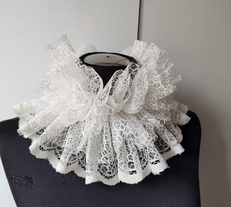 K&ouml;nnte beinhalten: Ein wei&szlig;er Spitzenkragen mit mehreren Lagen aus zarter, durchbrochener Spitze. Der kreisf&ouml;rmige Kragen wird auf einer schwarzen Schaufensterpuppe pr&auml;sentiert, die das komplizierte Design und die Textur hervorhebt. Der Kragen ist ein historisches Modeaccessoire.