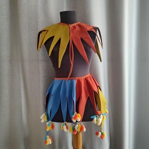 Peut inclure: Costume de bouffon coloré présenté sur un mannequin. Le costume comprend un col et une jupe jaunes, oranges et bleus avec des bords en pointe. La jupe est ornée de petits pompons décoratifs. Parfait pour une représentation théâtrale ou une soirée costumée.