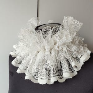 K&ouml;nnte beinhalten: Ein wei&szlig;er Spitzenkragen mit einem geschichteten, ger&uuml;schten Design. Der Kragen ist kreisf&ouml;rmig, mit einem schwarzen Innenraum, und wird auf einer schwarzen Schaufensterpuppe pr&auml;sentiert. Die Spitze hat ein zartes, offenes Webmuster und erzeugt ein strukturiertes Aussehen.