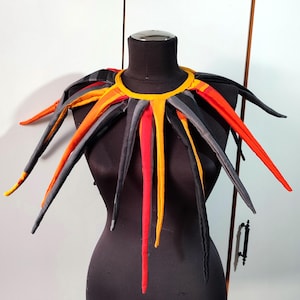 May include: A fabric collar accessory featuring a circular yellow neckline with multiple radiating, pointed fabric spikes in alternating shades of black, dark gray, bright orange, and red, extending outward from the center in a sunburst pattern.