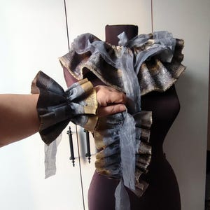May include: A pair of gray and gold ruffled fabric cuffs with a long gray ribbon. The cuffs are worn on a person's arm.
