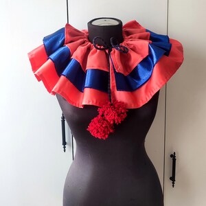 May include: A layered, ruffled collar in coral and blue satin, adorned with a black tie and two red pom-poms. The collar is displayed on a dark mannequin, showcasing its theatrical design. This costume accessory adds a touch of flair.