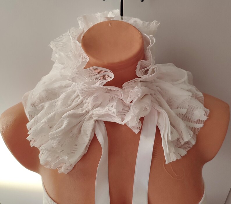 K&ouml;nnte beinhalten: Ein wei&szlig;er, ger&uuml;schter Kragen aus zartem Stoff, gebunden mit wei&szlig;en B&auml;ndern. Der Kragen wird auf einer Schaufensterpuppe pr&auml;sentiert, um seine Textur und sein Design hervorzuheben. Es ist ein Modeaccessoire.