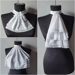 May include: Three white ruffled fabric collars, each with a different style and shape. The collars are displayed on mannequins with black fabric bodies.