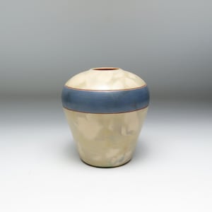 Puede incluir: Un jarrón de cerámica con cuerpo color crema y una ancha banda horizontal azul en el centro. El borde superior es de color marrón rojizo. El jarrón tiene forma redondeada y una pequeña abertura en la parte superior.