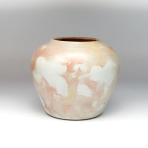 May include: A white ceramic vase with a light pink glaze and a mottled pattern. The vase has a rounded body and a wide opening.