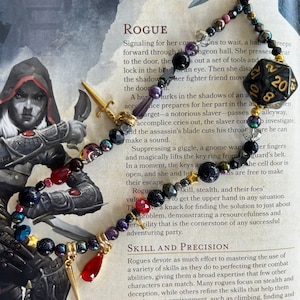 May include: A close-up of a necklace with black, red, gold, and purple beads. The necklace has a gold dagger charm, a gold star charm, and a red teardrop charm. The necklace is on a book with text about the Rogue class in a role-playing game. The text reads "Signaling for her cons to wait, a halm reeps forward through the dungeon hall. She presses..." and "SKILL AND PRECISION".