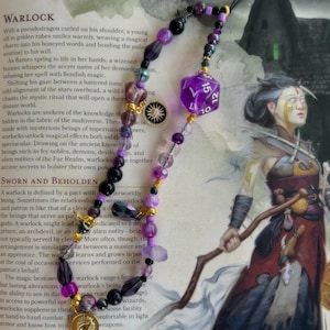 May include: A purple and gold beaded necklace with a purple 20-sided die charm and a gold sun charm. The necklace is on a book with text about warlocks in a fantasy role-playing game.