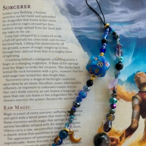 May include: A blue and gold 20-sided die charm with a crescent moon charm and various beads in shades of blue, purple, and black. The charm is attached to a black cord. The text "SORCERER" and "RAW MAGIC" are visible on the page behind the charm.