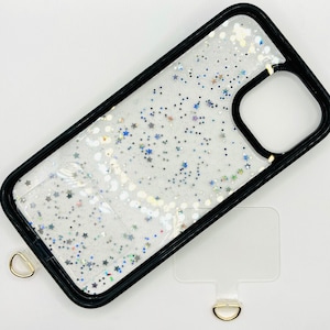 May include: A clear phone case with a black border and a glittery design featuring silver and blue stars. The case has a gold ring attached to the bottom.