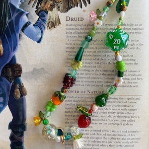 May include: A close-up of a green 20-sided die with a white background and black numbers. The die is surrounded by a colorful beaded necklace with charms, including flowers, leaves, and berries. The necklace is resting on a page of a book about druids, with text about their powers and connection to nature.
