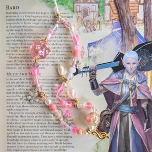 May include: A pink and white beaded necklace with a gold treble clef charm. The necklace is lying on a book with text about the role of music and magic in a fantasy world. The text includes the words "Music and Magic".