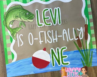 Puede incluir: Una pancarta pintada a mano con temática de pesca. La pancarta presenta un pez verde, un flotador rojo y blanco, y las palabras "LEVI IS O-FISH-ALLY ONE" en letras verdes y blancas. El fondo incluye agua, juncos y un borde verde y marrón.