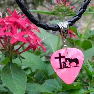 May include: A pink guitar pick necklace with a black leather cord. The pick features a silhouette of a person kneeling in prayer with a cross and a horse.