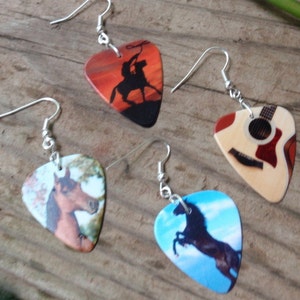 Puede incluir: Cuatro pendientes de p&uacute;as de guitarra con ganchos plateados. Una p&uacute;a presenta a un vaquero lazando un toro al atardecer. Otra representa una guitarra. Las otras dos muestran caballos, uno marr&oacute;n y otro negro, contra un cielo azul.