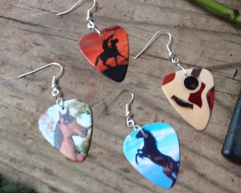 Puede incluir: Cuatro pendientes de p&uacute;as de guitarra con ganchos plateados. Una p&uacute;a presenta a un vaquero lazando un toro al atardecer. Otra representa una guitarra. Las otras dos muestran caballos, uno marr&oacute;n y otro negro, contra un cielo azul.