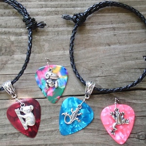 May include: Four guitar pick necklaces with silver animal charms: a koala, a frog, a lizard, and a frog. The picks are red, multicolored, blue, and pink, attached to black braided cords. The background is a weathered wooden surface.