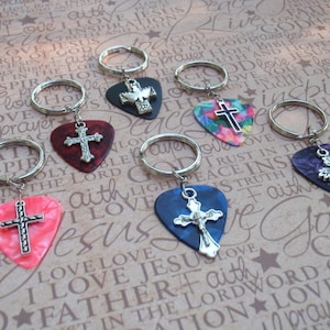 May include: Six guitar picks with silver cross charms attached to key rings. The picks are in various colors, including red, blue, purple, and pink. The crosses are all the same design, with a silver finish.